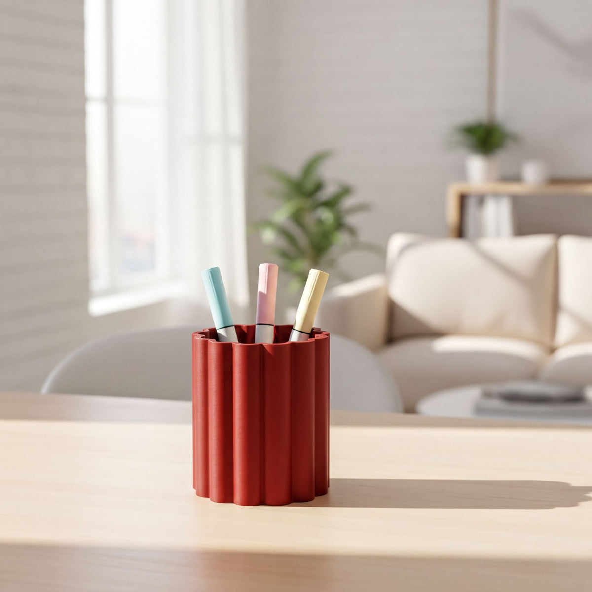 A red cylindrical container holding three colored markers is placed on a wooden table in a bright, well-lit room with a white sofa and a plant in the background.