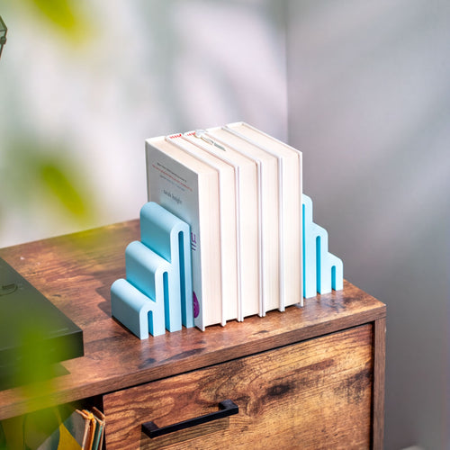 A set of light blue and white geometric bookends holding a row of books on top of a rustic wooden cabinet, with soft leaf shadows on a light gray wall in the background.