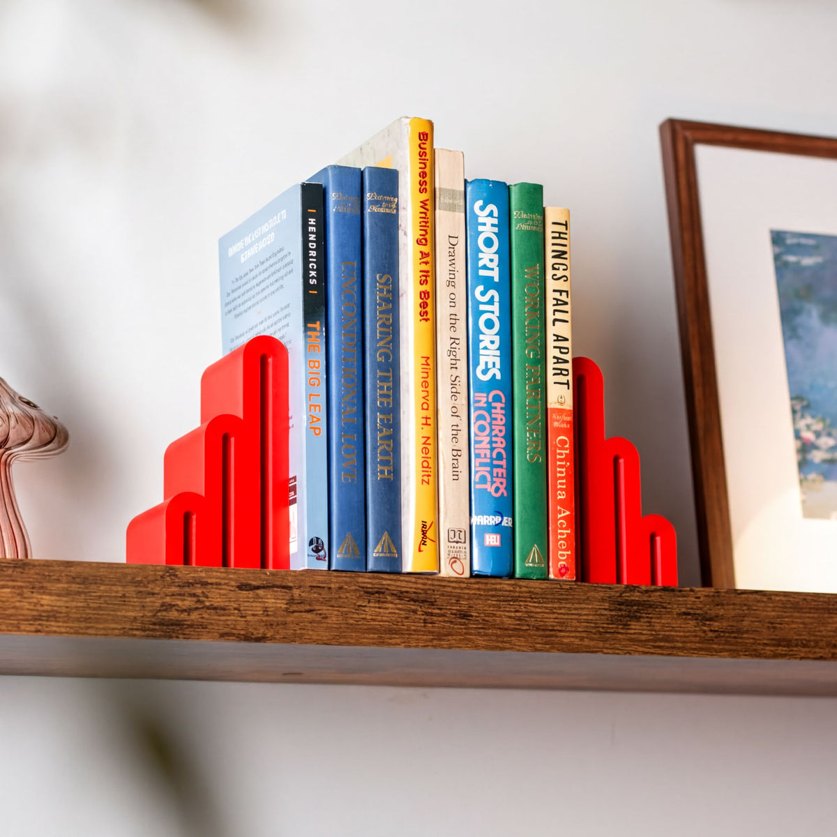 Red geometric bookends holding a row of colorful books on a wooden shelf against a white wall.