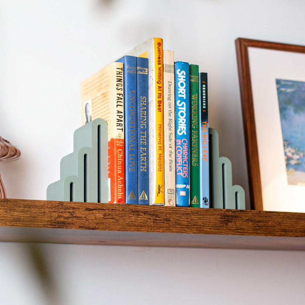 A set of modern, scalloped-edge bookends in a muted sage green color holding a row of colorful books on a wooden shelf against a white wall.