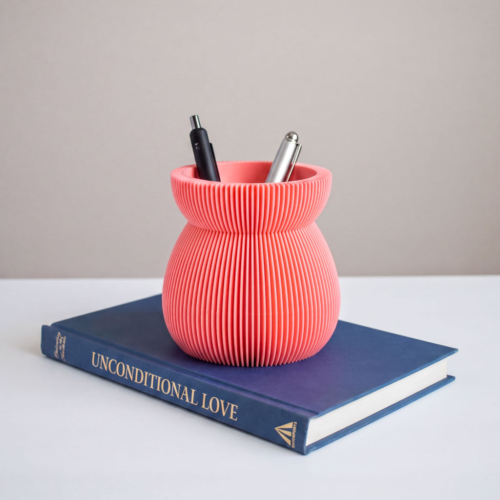 A pink, ribbed vase holding pens and a blue book titled "UNCONDITIONAL LOVE" is placed on a white surface.