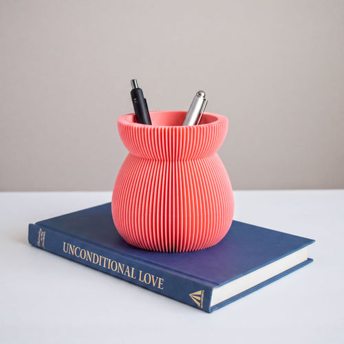 A pink, ribbed vase holding pens and a blue book titled "UNCONDITIONAL LOVE" is placed on a white surface.