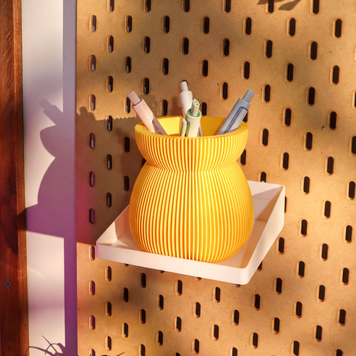 A yellow, ribbed container holding various colored pens is displayed on a pegboard against a wooden wall.