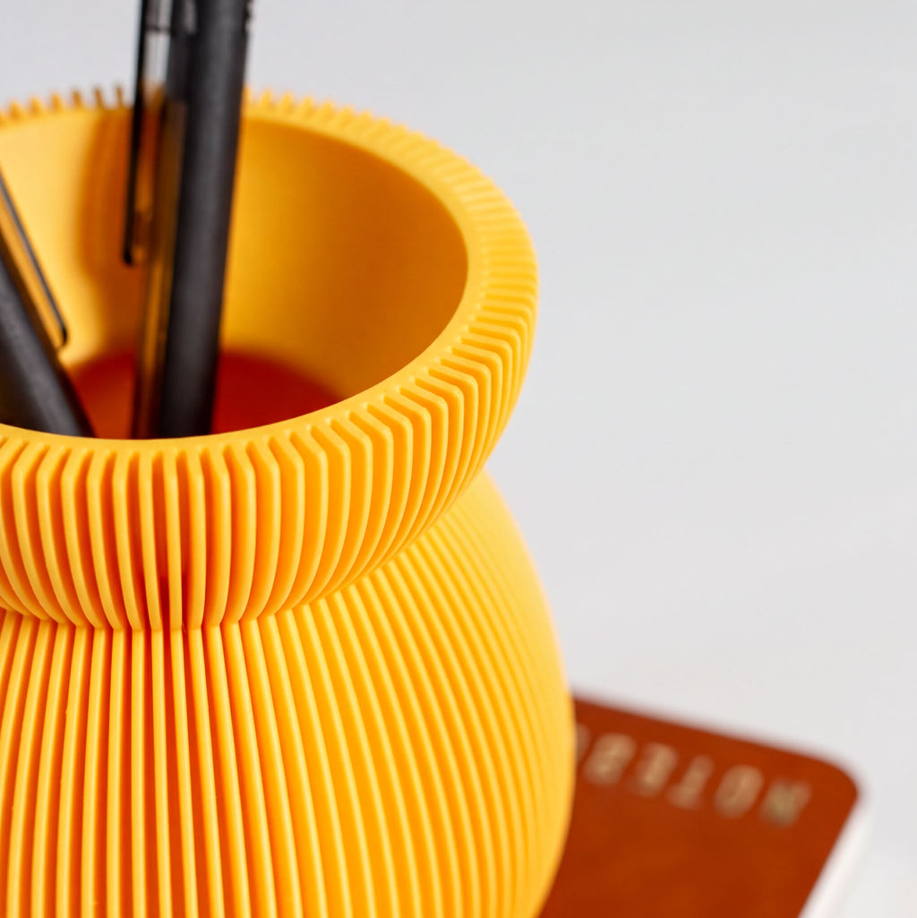 A close-up view of a yellow, textured vase or container holding pens and pencils, with a blurred background.