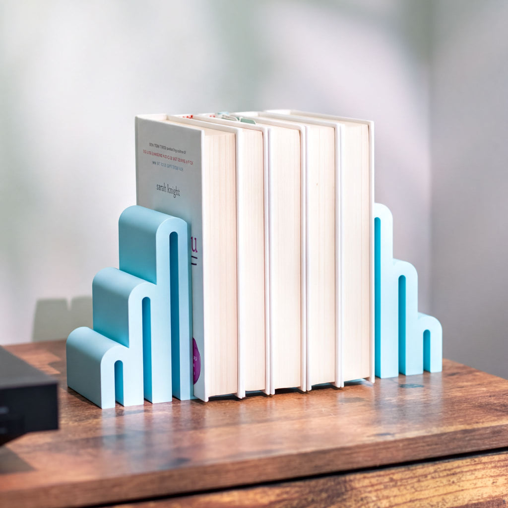 A set of modern step-style bookends in light blue and turquoise holding a row of books on a wooden table, with a softly blurred indoor background.