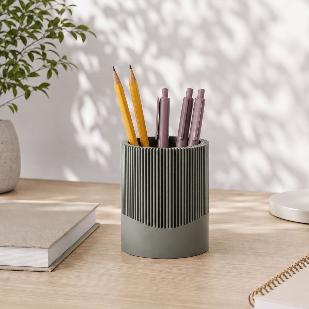 A gray cylindrical pencil holder filled with various colored pencils and pens sits on a wooden desk, accompanied by a book and a plant.